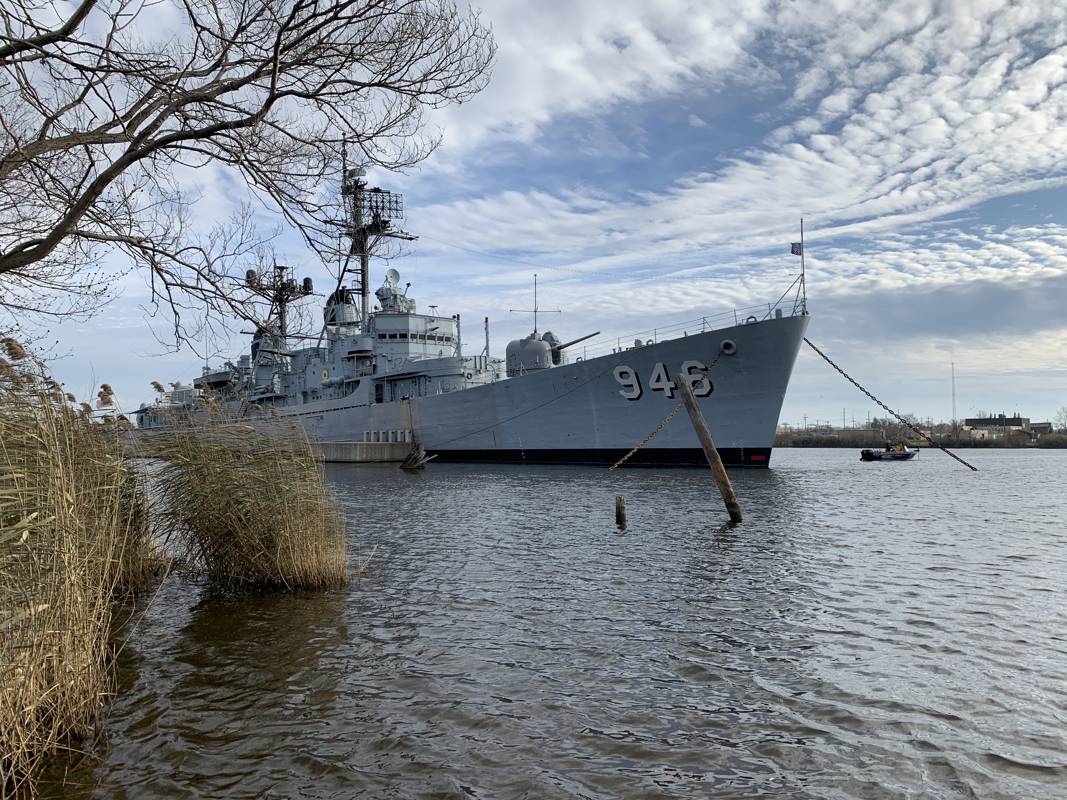 Bow view of destroyer Edson taken from the shore, a grey ship with number 946 on the bow in the Saginaw River with a marsh and tree to the left.