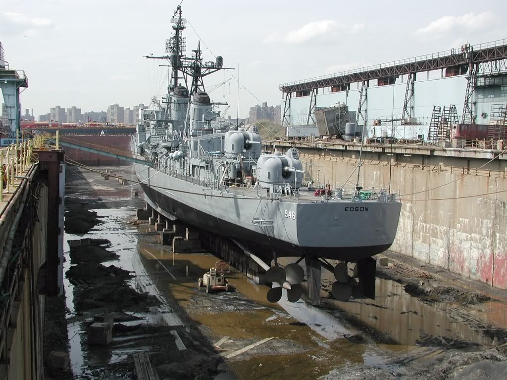 Stern view of a large grey destroyer sitting on keel blocks in the bottom of a concrete dry dock with a large blue, windowless building on the right.