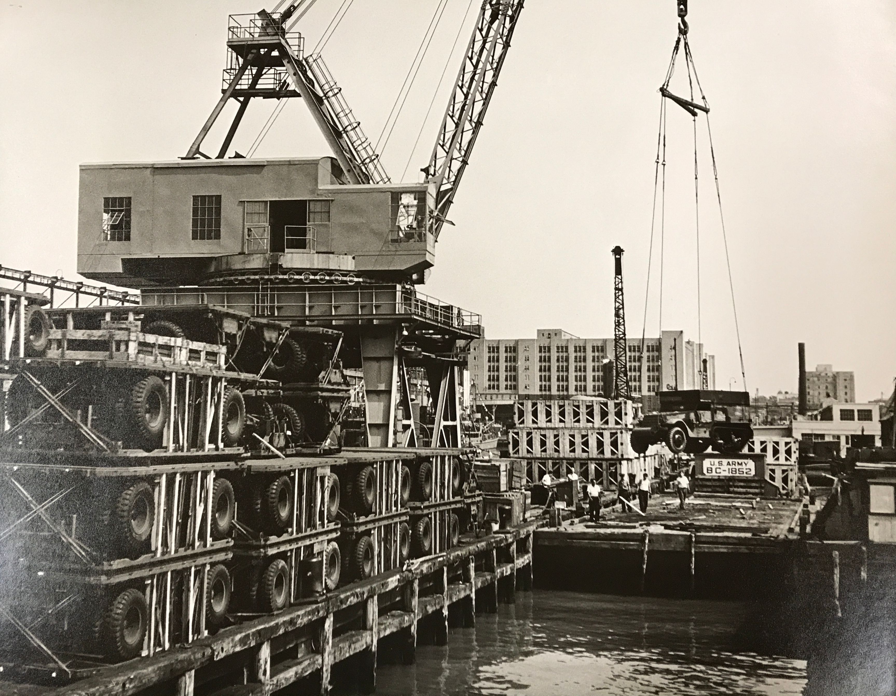A crane lifting a jeep onto a barge with the warehouse of the Brooklyn Army Terminal visible in the background.