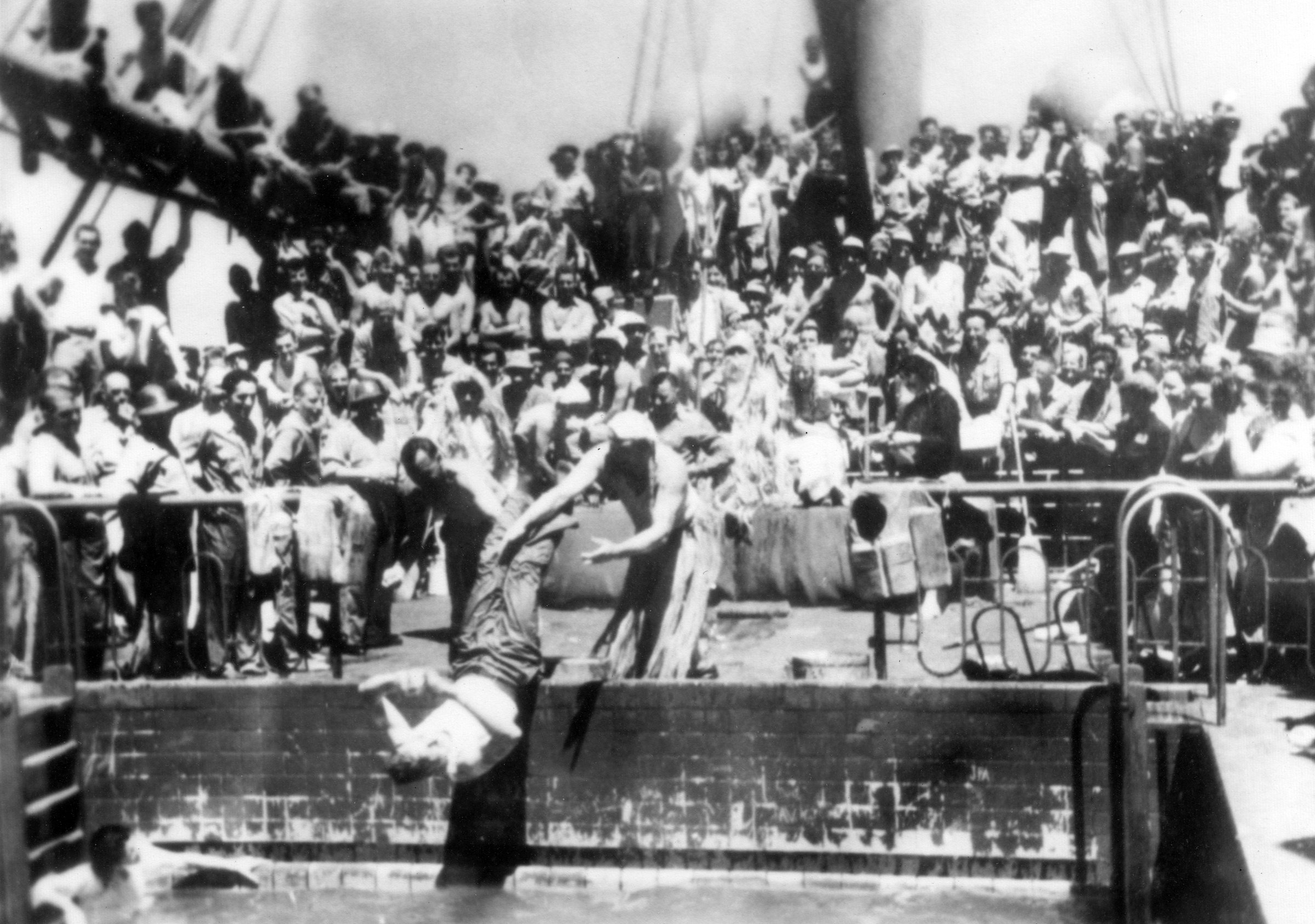 Black and white photo of hundreds of soldiers on the deck of a ship watching another soldier being thrown into a swimming pool.