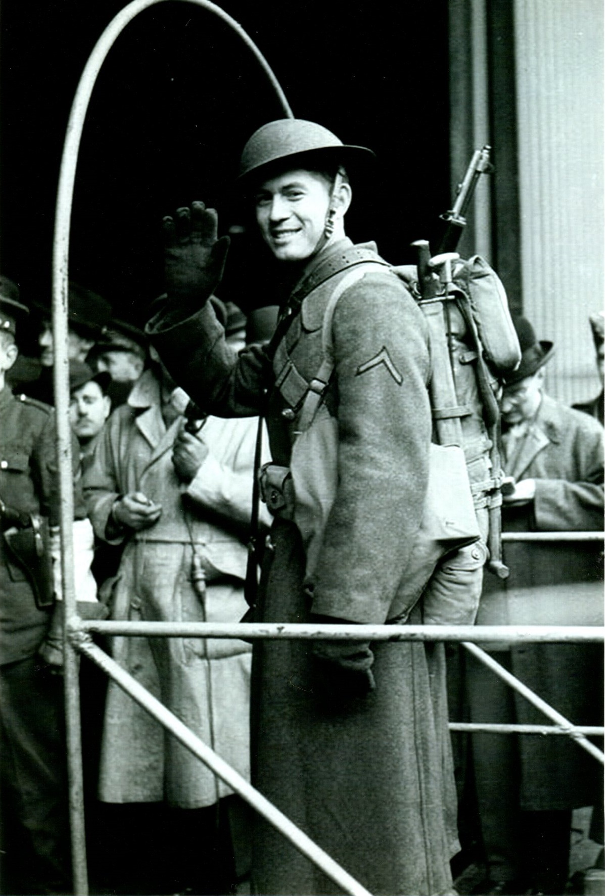 Black and white photo of a soldier waving from the gangplank of a ship with soldiers in the background.