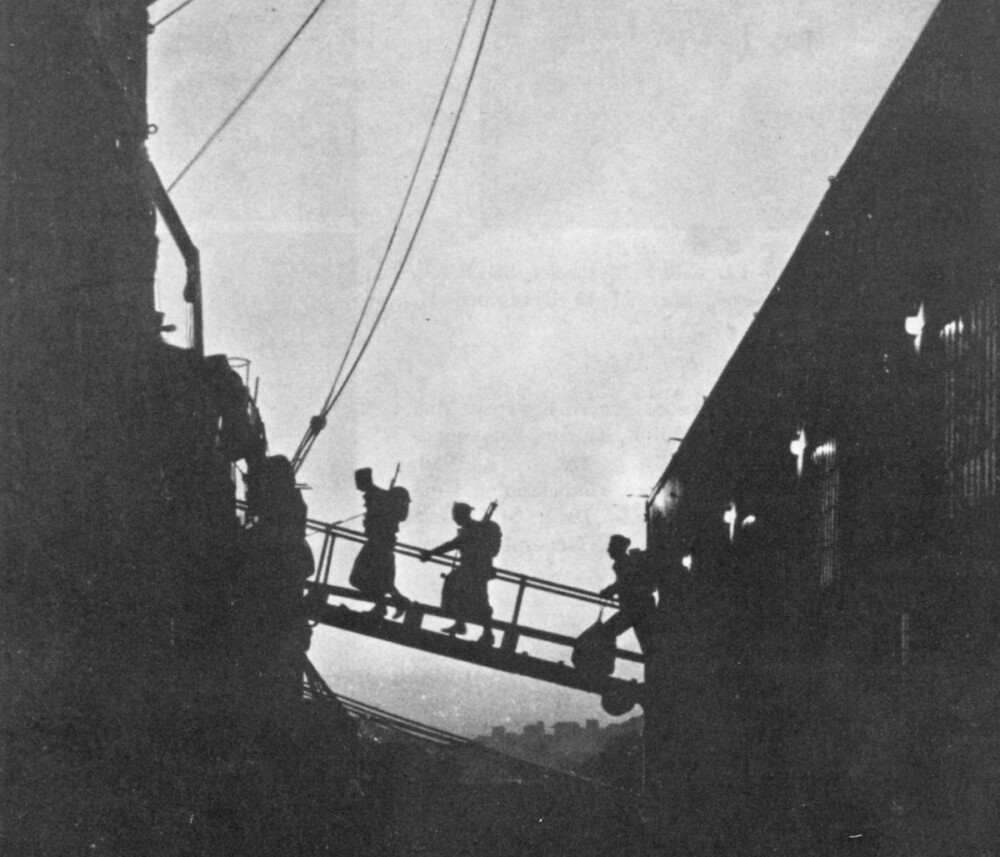 Black and white photo of two soldiers walking up a gangplank onto a ship at dusk.