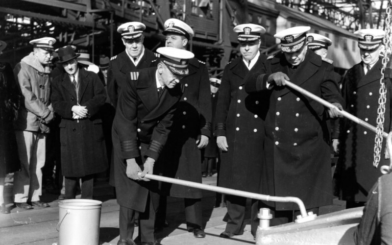 Black and white photo of five naval officers standing on a large metal plate, one of them turning a long handle attached to a riveting machine.