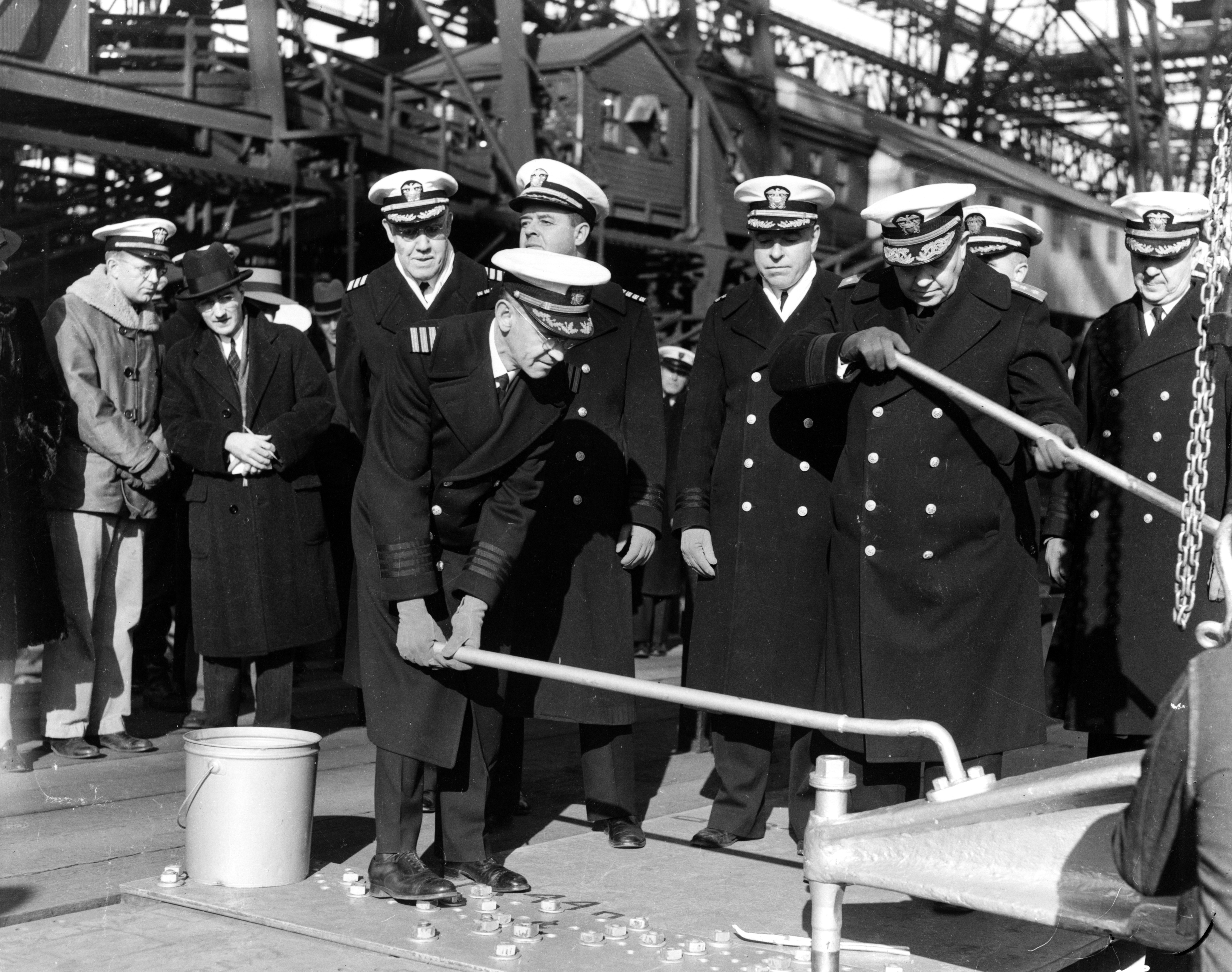 Black and white photo of five naval officers standing on a large metal plate, one of them turning a long handle attached to a riveting machine.