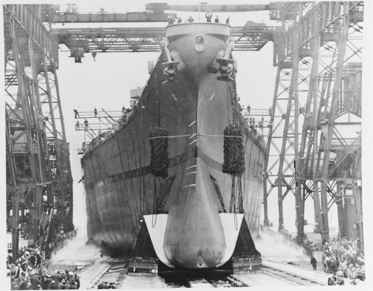 Black and white photo of a battleship sliding down a ramp into the East River, with people standing on the bow waving.