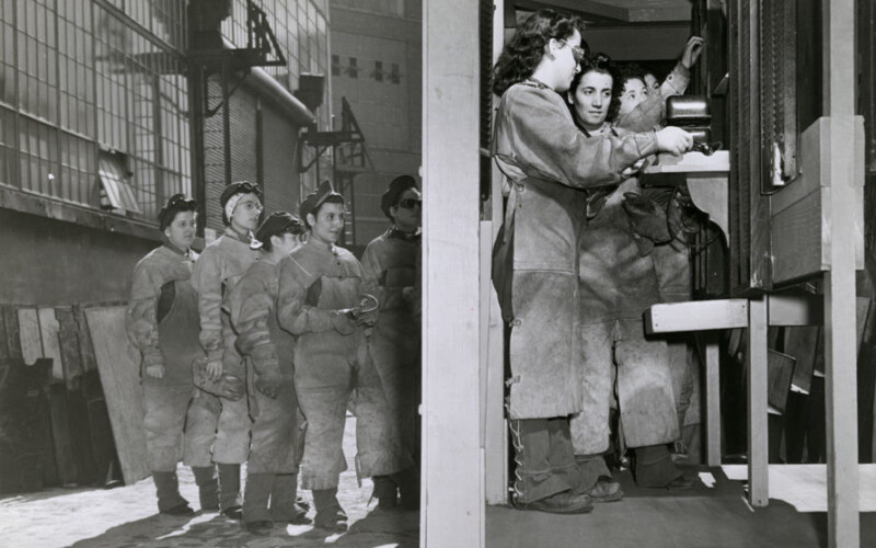 8-10 female workers lining up at the Brooklyn Navy Yard to punch in wearing heavy smocks for welding.
