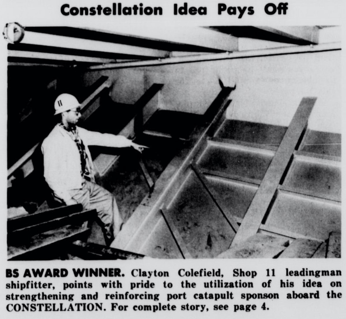 Black and white newspaper photo of a worker wearing a helmet standing and pointing inside a ship.