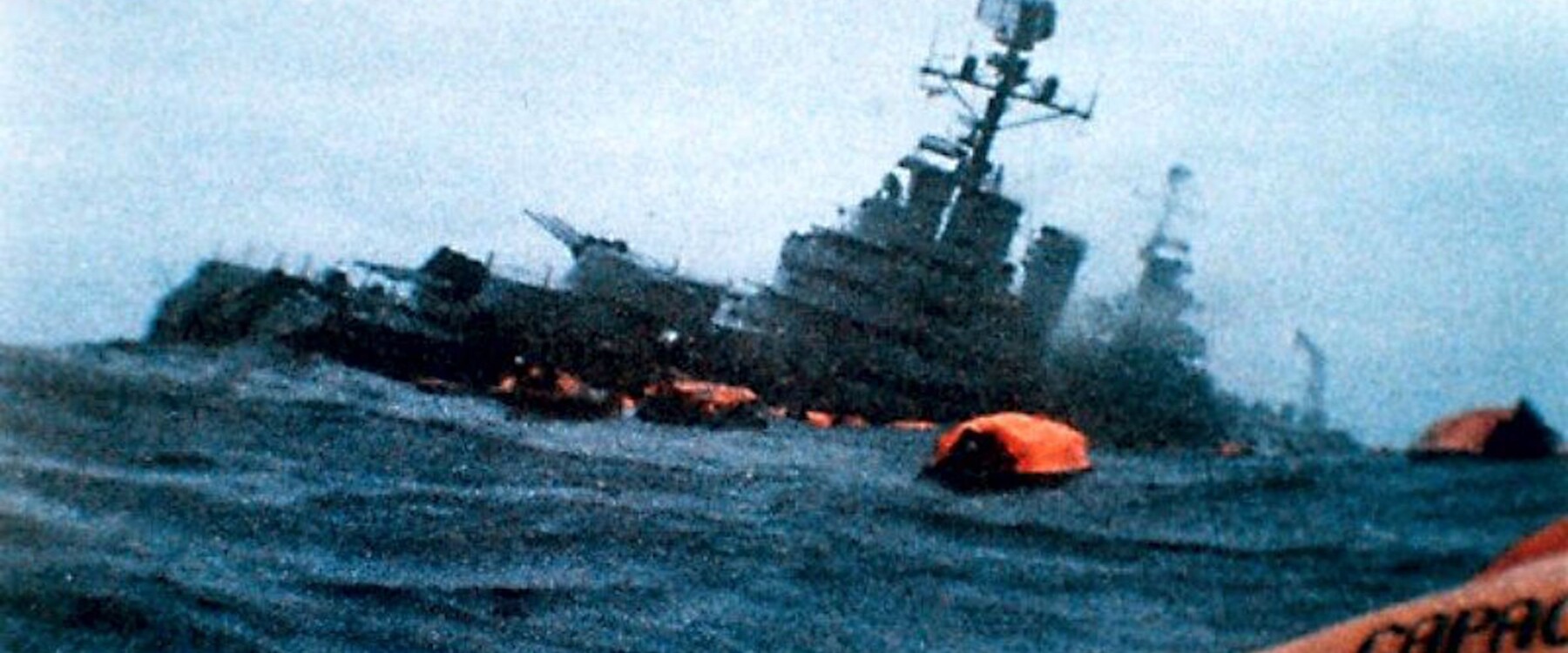 Color photo of a cruiser sinking with ocean in foreground and orange lifeboats on the water.