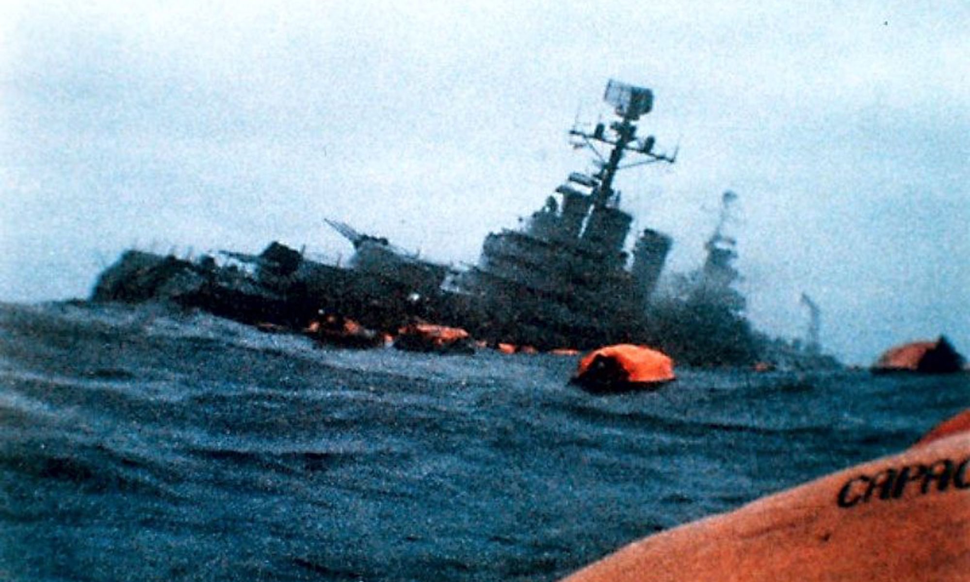 Color photo of a cruiser sinking with ocean in foreground and orange lifeboats on the water.