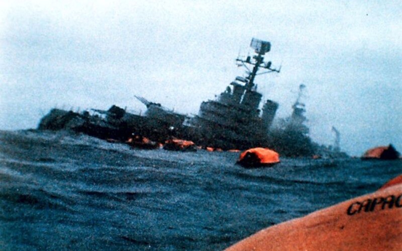 Color photo of a cruiser sinking with ocean in foreground and orange lifeboats on the water.