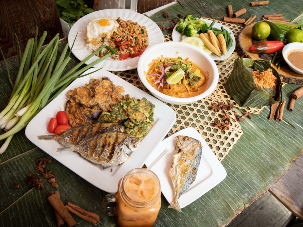 Photo of plates of food on a table, including fish, curry, carrots, rice, and Thai iced tea