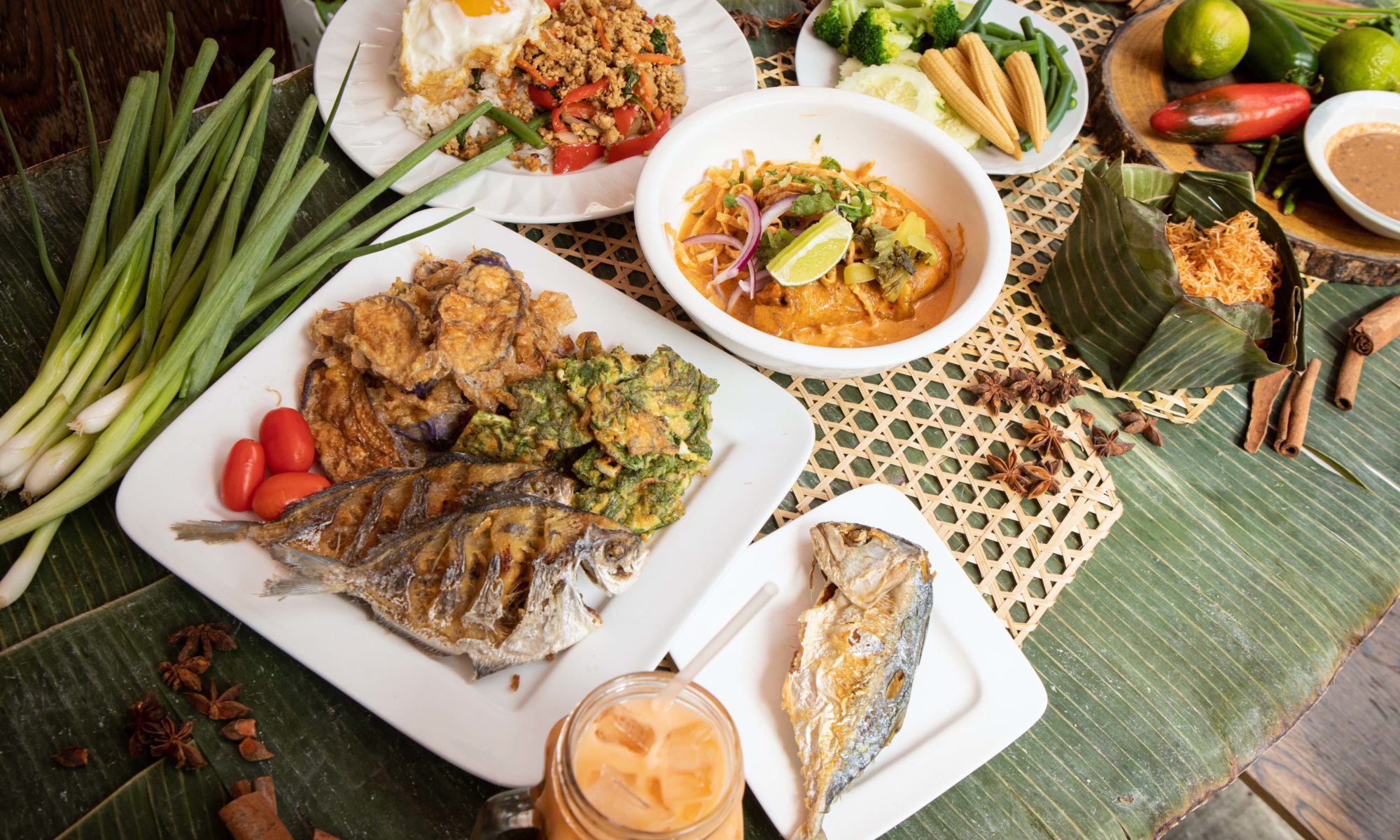 Photo of plates of food on a table, including fish, curry, carrots, rice, and Thai iced tea