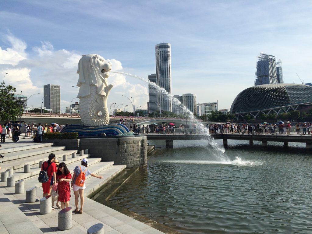 Photo of the Singapore Merlion statue spraying water into a pool with people watching and city skyline in the distance