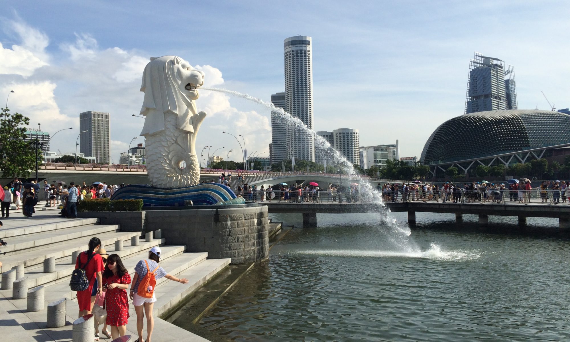 Photo of the Singapore Merlion statue spraying water into a pool with people watching and city skyline in the distance