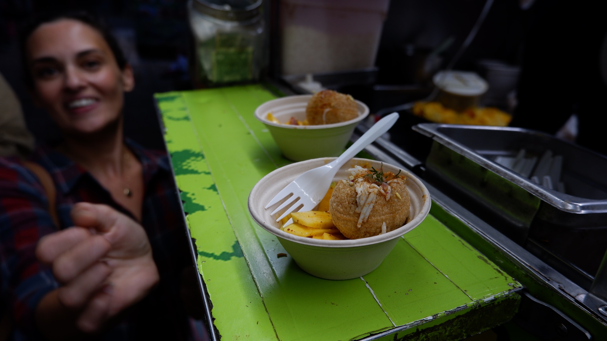 A small bowl filled with Bangladeshi fushka and a fork sits on a green shelf attached to a food cart, with a woman reaching for the bowl.