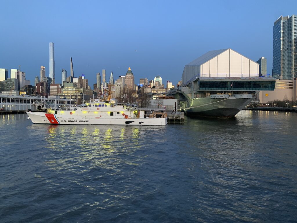 Photo of a white Coast Guard vessel with a red stripe near its bow and the words "US Coast Guard." It is in the Hudson River and illuminated with yellowish lights, with the Manhattan skyline behind it. USS Intrepid sits to the right of the photo.