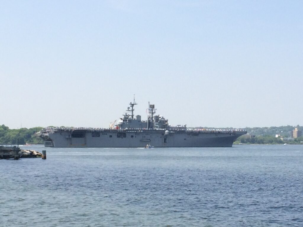 Photo of USS Wasp, an amphibious assault ship that resembles an aircraft carrier with a large, flat flight deck. The crew is standing out on the deck as the ship sails into New York Harbor.