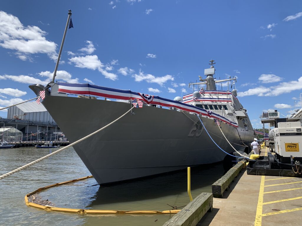 Photo of a grey-hulled Navy ship, USS Cooperstown, with a view from the pier facing towards the bow. red, white, and blue bunting is stretched around the ship's gunwhales and superstructure. 
