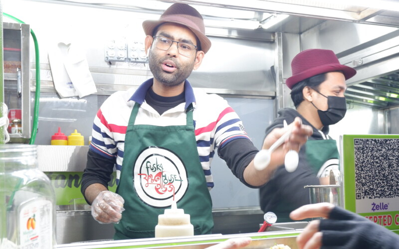 Vendor from Fushka House cart wearing a green apron and brown hat passes out bowls of food.