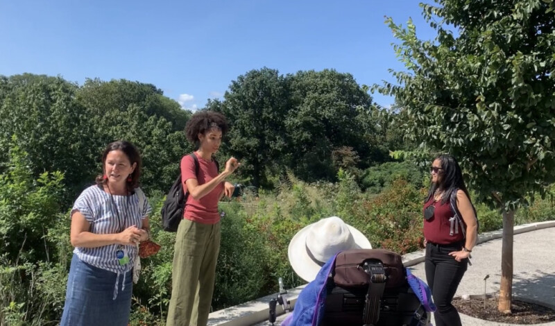 A woman speaks outside on a pathway in front of trees and bushes as an ASL interpreter signs and two others look at them